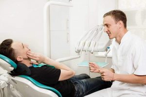 Dentist talking to patient who is in a dental chair and holding his jaw as though he’s in pain.